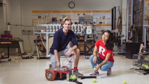 Male and female students kneeling in a lab with a device that collects tennis balls. Male has blond hair and is wearing shorts with a navy blue shirt. Female on right had black hair and is wearing a red t-shirt with a white letter R and jeans.