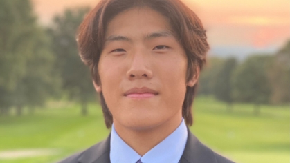 Asian male college student poses outdoors for a headshot. He is wearing a dark suit, light blue shirt and blue tie.