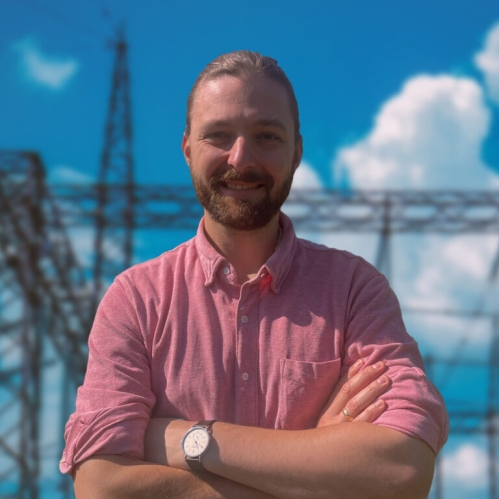 head shot of male with arms folded in front of a power grid with short blonde hair and moustache and beard, wearing a salmon colored button down shirt