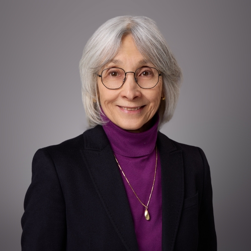 Headshot of female with short grey hair with bangs and eyeglasses wearing a purple turtleneck and black jacket.