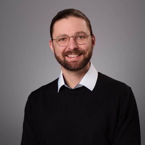 Headshot of male with short dark brown hair, moustache, beard and eyeglasses wearing a white collared shirt beneath a black pullover with a