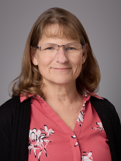 Headshot of female with light brown shoulder length hair, bangs, and eyeglasses wearing a red patterned blouse and a black sweater.