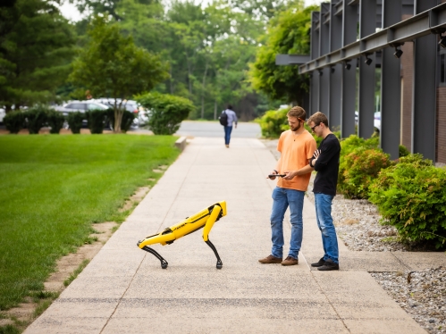 Two male students work with a yellow robotic dog outdoors on a sidewalk.