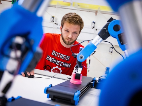 Graduate student wearing a red T-shirt works in a lab with robotic arms lifting small objects.