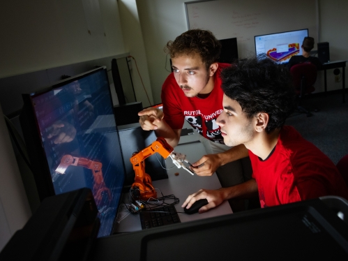 two male students wearng red shirts looking at computer screen with a small robotic arm on the desk in front of them 