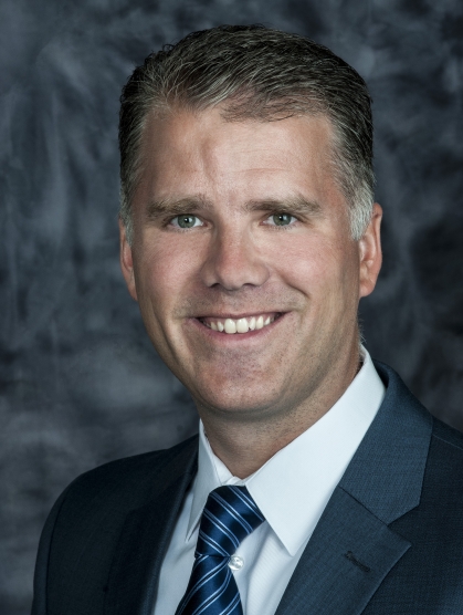 Head shot of white male wearing a dark suit with a white shirt and blue tie.