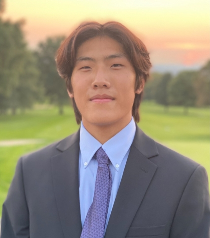 Asian male college student poses outdoors for a headshot. He is wearing a dark suit, light blue shirt and blue tie.