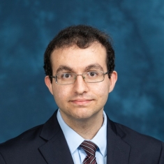 Headshot of male with short dark brown hair with eyeglasses wearing a black suit, white shirt and patterned tie.